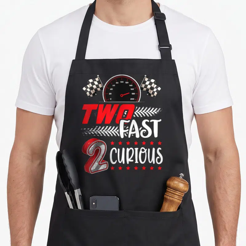 Fast racing apron - Speedometer flanked by checkered flags above stacked 'TWO FAST CURIOUS' text with red number 2 — shown on a black cotton canvas apron against a white background