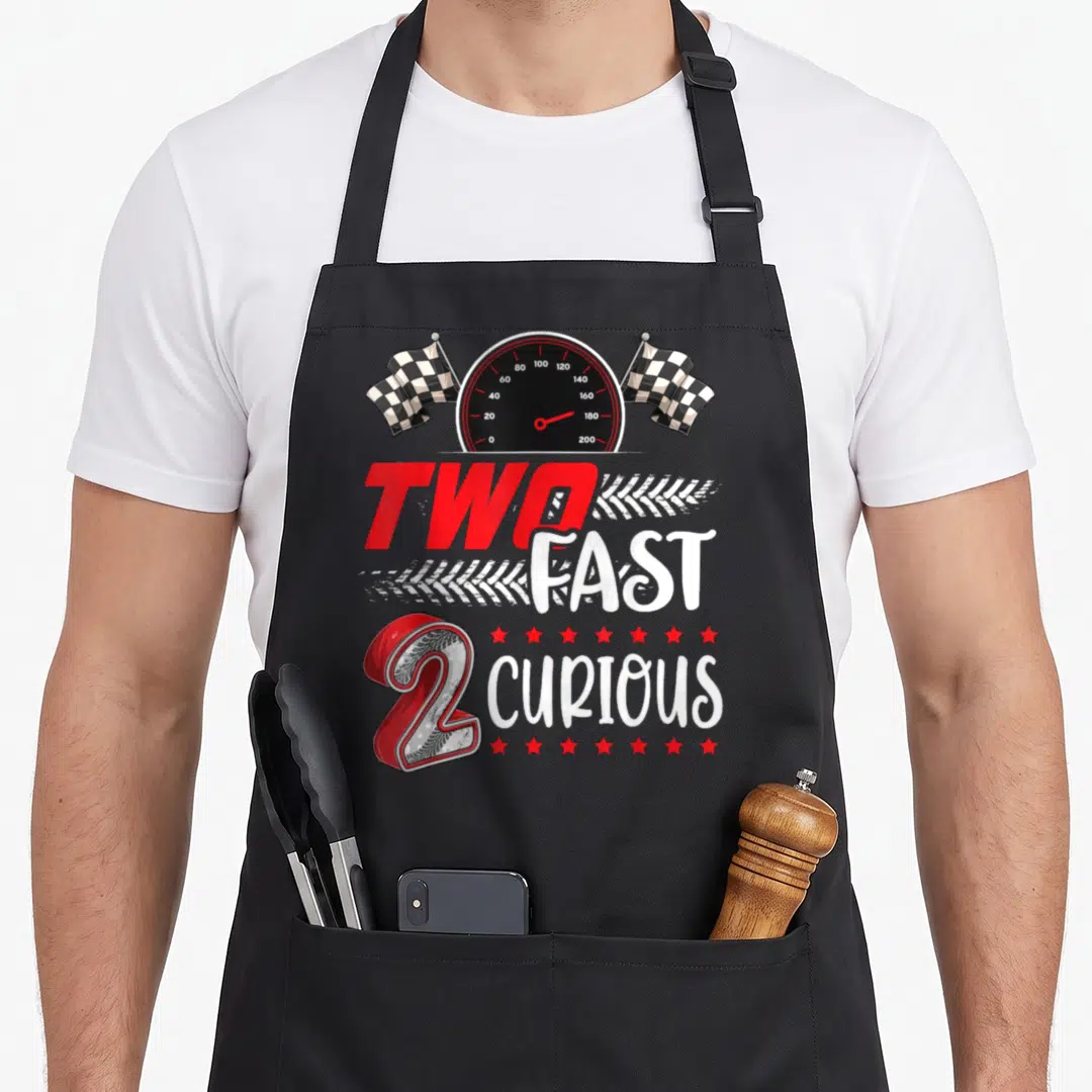 Fast racing apron - Speedometer flanked by checkered flags above stacked 'TWO FAST CURIOUS' text with red number 2 — shown on a black cotton canvas apron against a white background