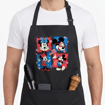 Grid 4th of july apron - Mickey and Minnie in 2x2 grid with red white blue splatter, stars, fireworks, and flags — shown on a black cotton canvas apron against a white background