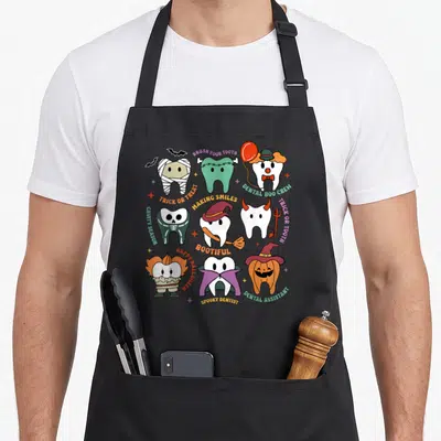 Boo dentist apron - Seven costumed cartoon teeth in orange, purple, green, and white surrounded by Halloween dental pun — shown on a black cotton canvas apron against a white background