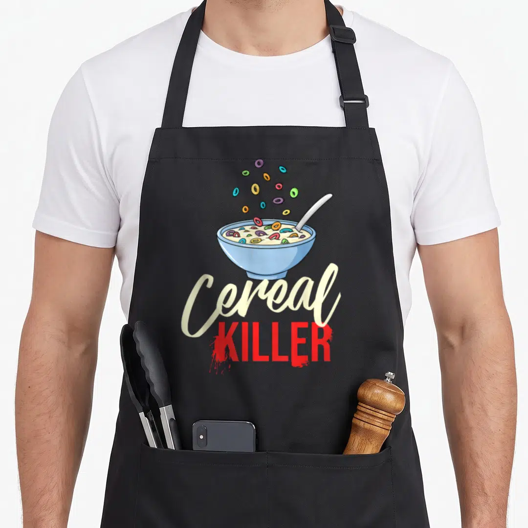 Cereal box apron - Cartoon blue bowl with floating loops and Cereal KILLER text in cream and red — shown on a black cotton canvas apron against a white background