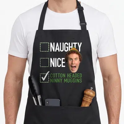 Naughty elf apron - Cartoon man head overlapping three checkboxes labeled naughty nice and cotton headed ninny muggins — shown on a black cotton canvas apron against a white background