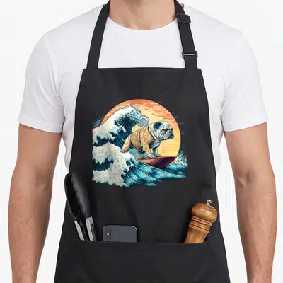 Surfing english chef aprons - Cartoon bulldog surfing blue wave inside orange sun circle — shown on a black cotton canvas apron against a white background