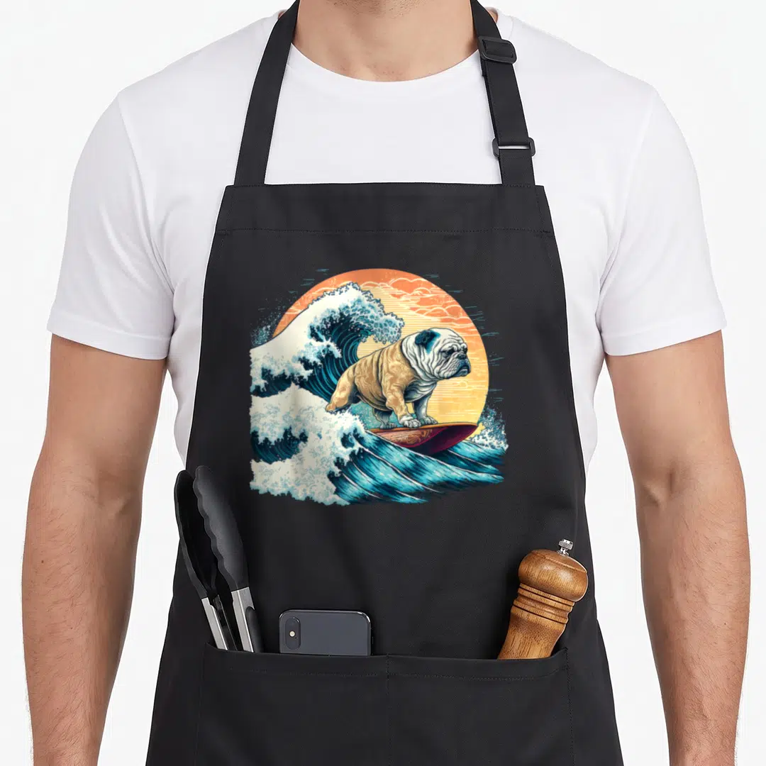 Surfing english chef aprons - Cartoon bulldog surfing blue wave inside orange sun circle — shown on a black cotton canvas apron against a white background