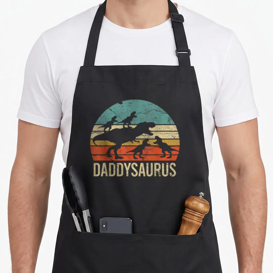 Dinosaur daddy aprons - Retro striped sun with T-Rex silhouettes and Daddysaurus text below — shown on a black cotton canvas apron against a white background