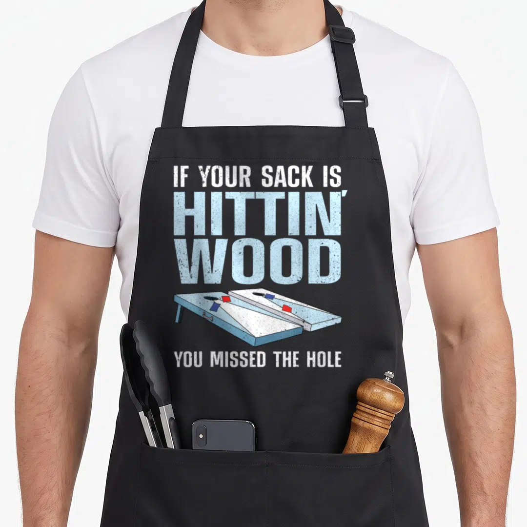 Cornhole bean bags and aprons - Text if your sack is hittin wood above blue white and red cartoon cornhole boards — shown on a black cotton canvas apron against a white background