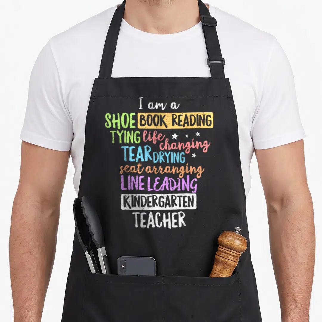 Teacher creative apron - Stacked colorful text listing teacher duties with three small white stars scattered among — shown on a black cotton canvas apron against a white background