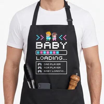 Baby pregnancy by company apron - Pixel characters blue pink loading bar DAD PLAYER MOM PLAYER text — shown on a black cotton canvas apron against a white background