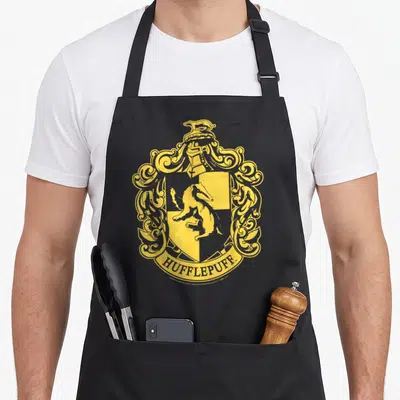 Hufflepuff simple apron - Yellow line-art crest with badger in shield, helmet, ornate mantling, and HUFFLEPUFF banner — shown on a black cotton canvas apron against a white background