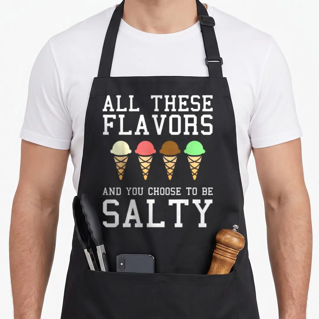 These meme apron - Stacked white text reading All These Flavors You Choose To Be Salty above four pink, brown, green — shown on a black cotton canvas apron against a white background