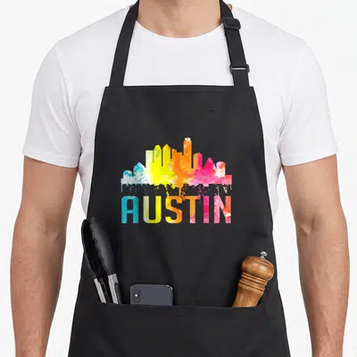 Watercolor texas aprons - AUSTIN text and skyline in blue yellow orange pink watercolor splashes — shown on a black cotton canvas apron against a white background