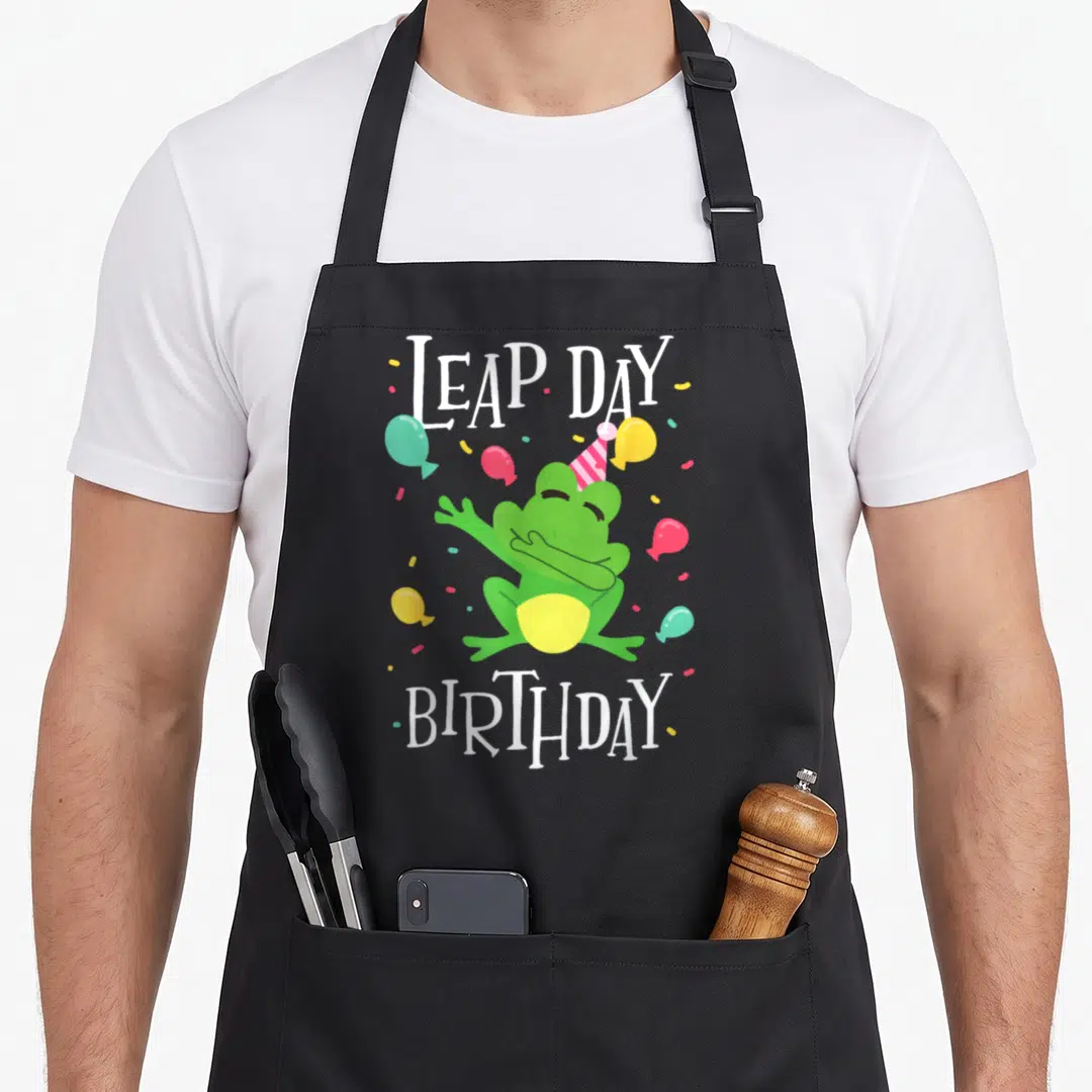 Frog birthday apron - Green frog dabbing wearing pink party hat with LEAP DAY BIRTHDAY text and balloons — shown on a black cotton canvas apron against a white background