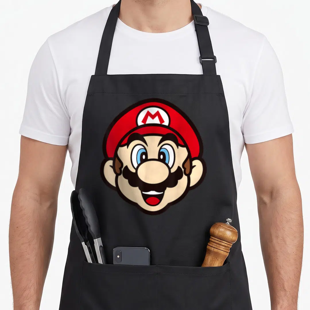 Music face apron - Cartoon Mario head with red hat and M logo in red white blue black and peach — shown on a black cotton canvas apron against a white background
