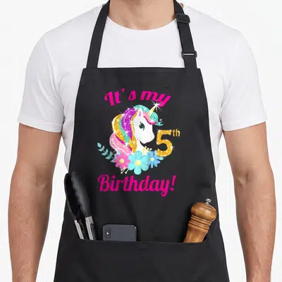 5th chef outfit apron - Unicorn head with pink mane, gold 5th, blue flowers, and It's my Birthday text — shown on a black cotton canvas apron against a white background