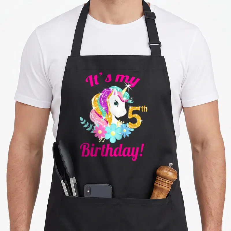 5th chef outfit apron - Unicorn head with pink mane, gold 5th, blue flowers, and It's my Birthday text — shown on a black cotton canvas apron against a white background