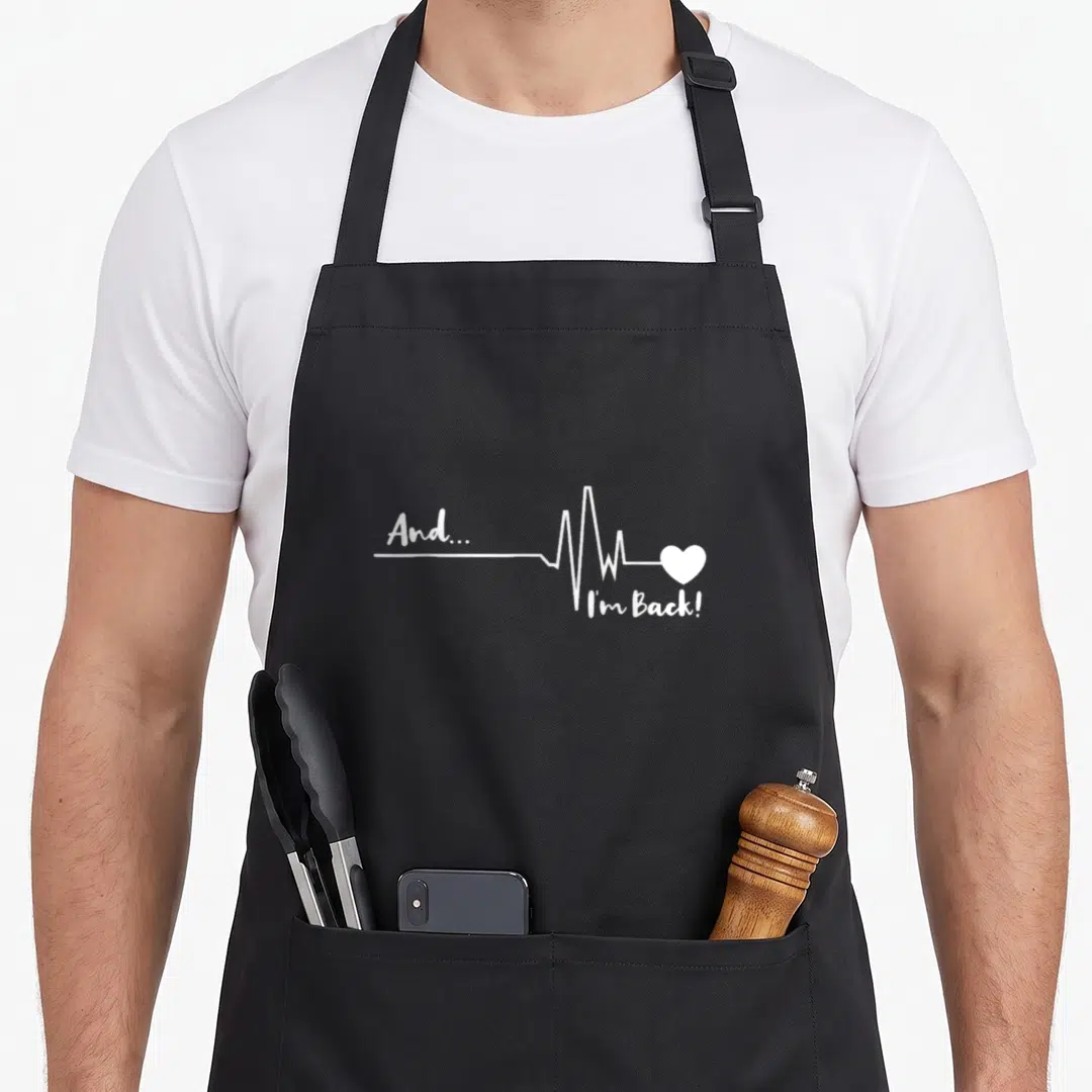 Surgery doctor apron - white heartbeat line with And... I'm Back! text and heart symbol — shown on a black cotton canvas apron against a white background