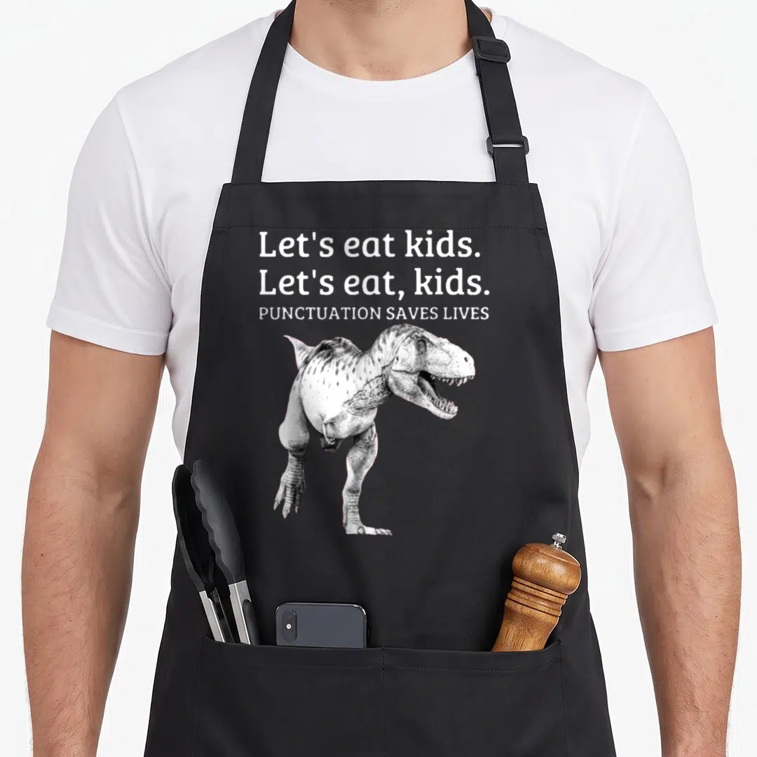 Punctuation let's eat apron - T-Rex illustration below three lines of white text about punctuation saving lives on grey — shown on a black cotton canvas apron against a white background