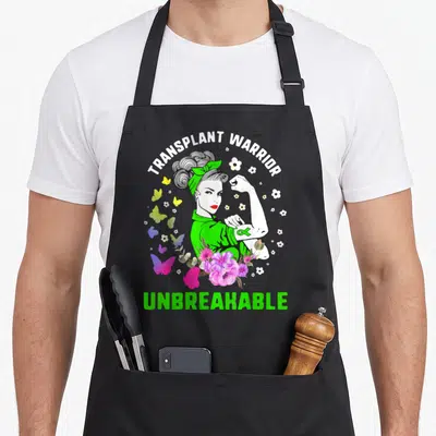 Unbreakable organ apron - Cartoon woman flexing arm with green ribbon, surrounded by butterflies and flowers, text — shown on a black cotton canvas apron against a white background