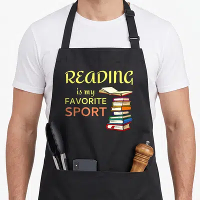 Sport reading books apron - Cartoon stack of multicolored books with one floating open volume and bold yellow — shown on a black cotton canvas apron against a white background