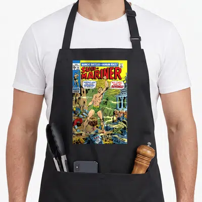 Namor bomber apron - Vintage comic cover with Namor, red title text, speech bubbles, and debris in primary colors — shown on a black cotton canvas apron against a white background