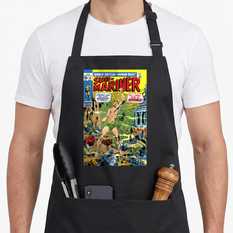 Namor bomber apron - Vintage comic cover with Namor, red title text, speech bubbles, and debris in primary colors — shown on a black cotton canvas apron against a white background