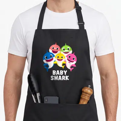 Pinkfong family aprons - Five cartoon sharks in blue, yellow, pink, red, green, white above BABY SHARK text — shown on a black cotton canvas apron against a white background