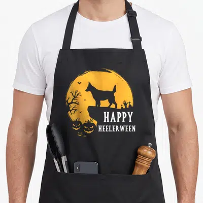 Moon dog kitchen apron - Grey heeler silhouette on orange moon with pumpkins and HAPPY HEELERWEEN text — shown on a black cotton canvas apron against a white background