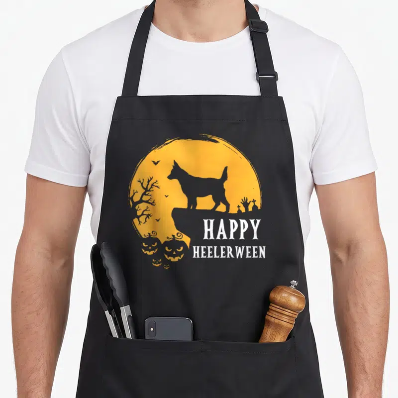 Moon dog kitchen apron - Grey heeler silhouette on orange moon with pumpkins and HAPPY HEELERWEEN text — shown on a black cotton canvas apron against a white background