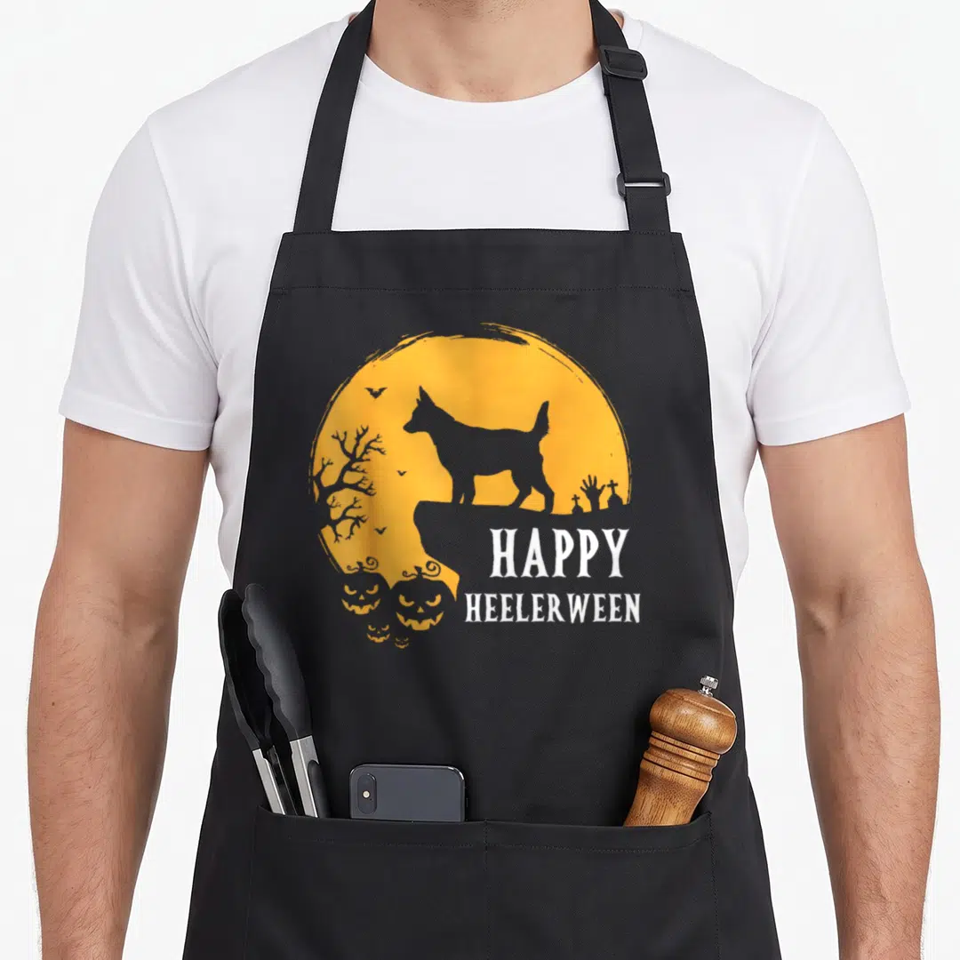 Moon dog kitchen apron - Grey heeler silhouette on orange moon with pumpkins and HAPPY HEELERWEEN text — shown on a black cotton canvas apron against a white background