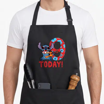 Tropical today birthday apron - Blue Stitch sitting with guitar in front of red floral number 9 and TODAY text — shown on a black cotton canvas apron against a white background
