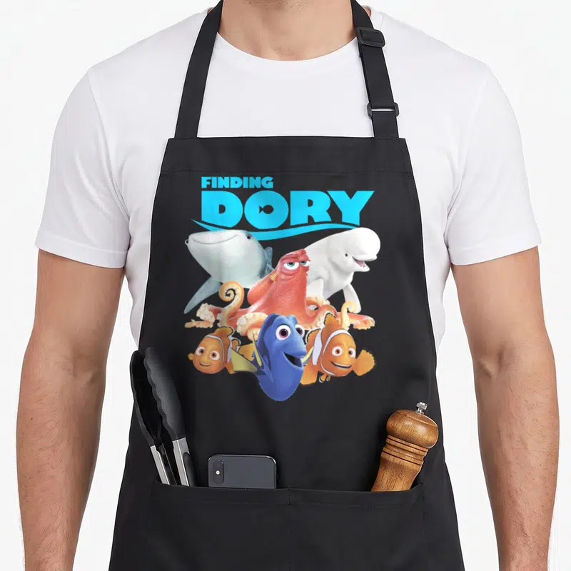 Finding crew apron - FINDING DORY text above blue tang, orange octopus, shark, beluga, and clownfish — shown on a black cotton canvas apron against a white background