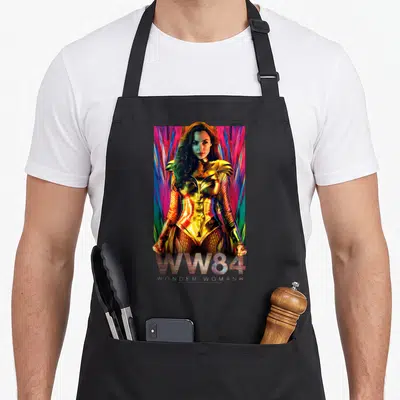 Warrior golden apron - Wonder Woman in gold armor with colorful rays and WW84 WONDER WOMANS text — shown on a black cotton canvas apron against a white background