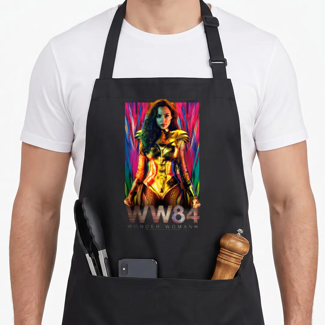 Warrior golden apron - Wonder Woman in gold armor with colorful rays and WW84 WONDER WOMANS text — shown on a black cotton canvas apron against a white background