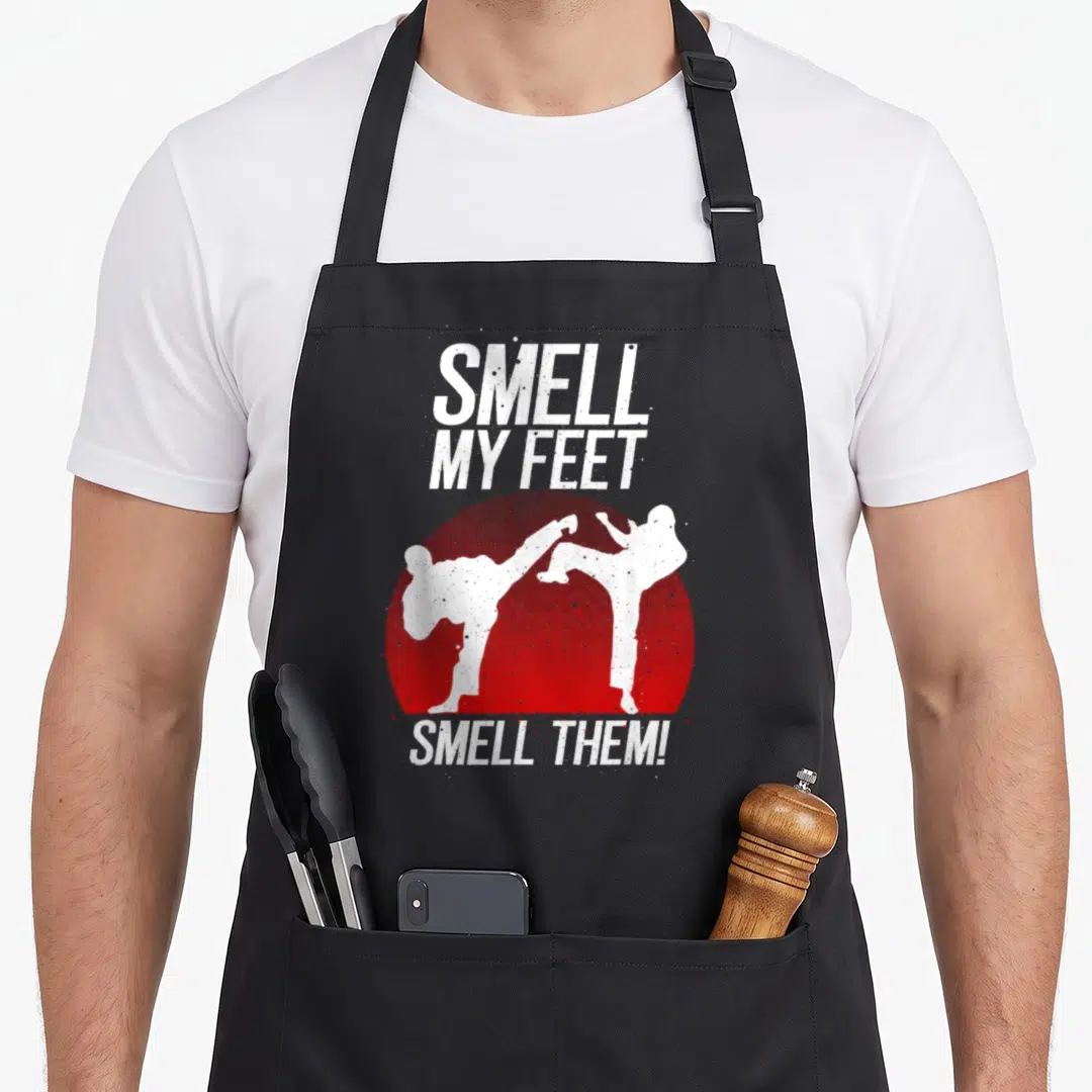 Smell karate apron - White SMELL MY FEET text above red circle with two white martial artist silhouettes fighting — shown on a black cotton canvas apron against a white background
