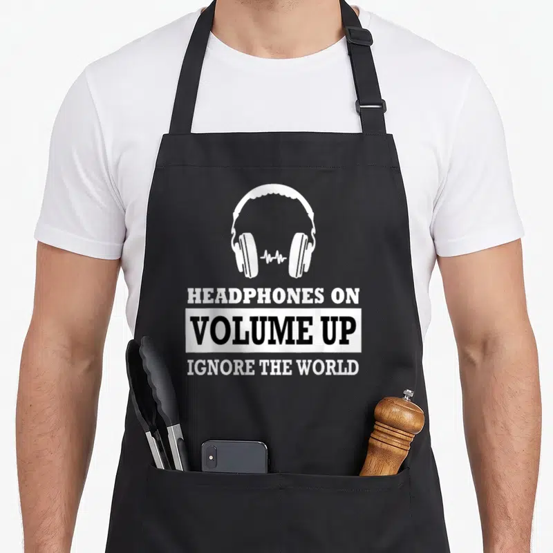 Volume headphones apron - White headphones graphic above stacked text reading HEADPHONES ON VOLUME UP IGNORE THE WORLD — shown on a black cotton canvas apron against a white background