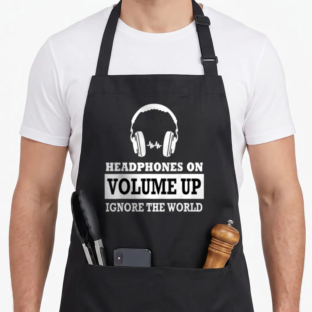 Volume headphones apron - White headphones graphic above stacked text reading HEADPHONES ON VOLUME UP IGNORE THE WORLD — shown on a black cotton canvas apron against a white background