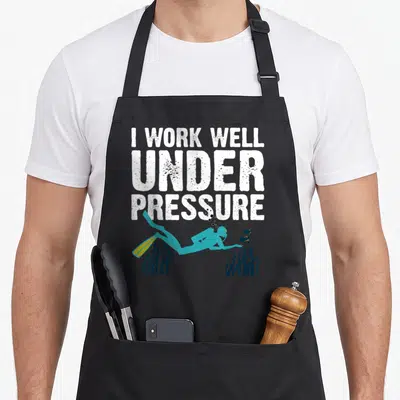 Diving snorkeling apron - Stacked text I work well under pressure above teal scuba diver and seaweed — shown on a black cotton canvas apron against a white background