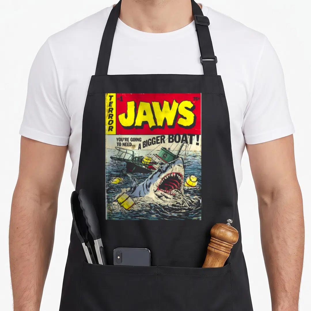 Pulp cover apron - Retro Jaws comic cover art with leaping shark and bold yellow lettering on red background — shown on a black cotton canvas apron against a white background