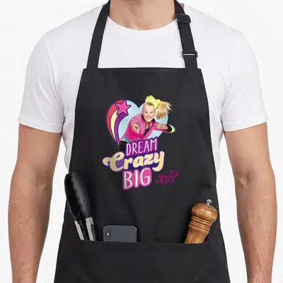 Dream shooting apron - Cartoon girl pointing with DREAM Crazy BIG text and shooting star heart — shown on a black cotton canvas apron against a white background