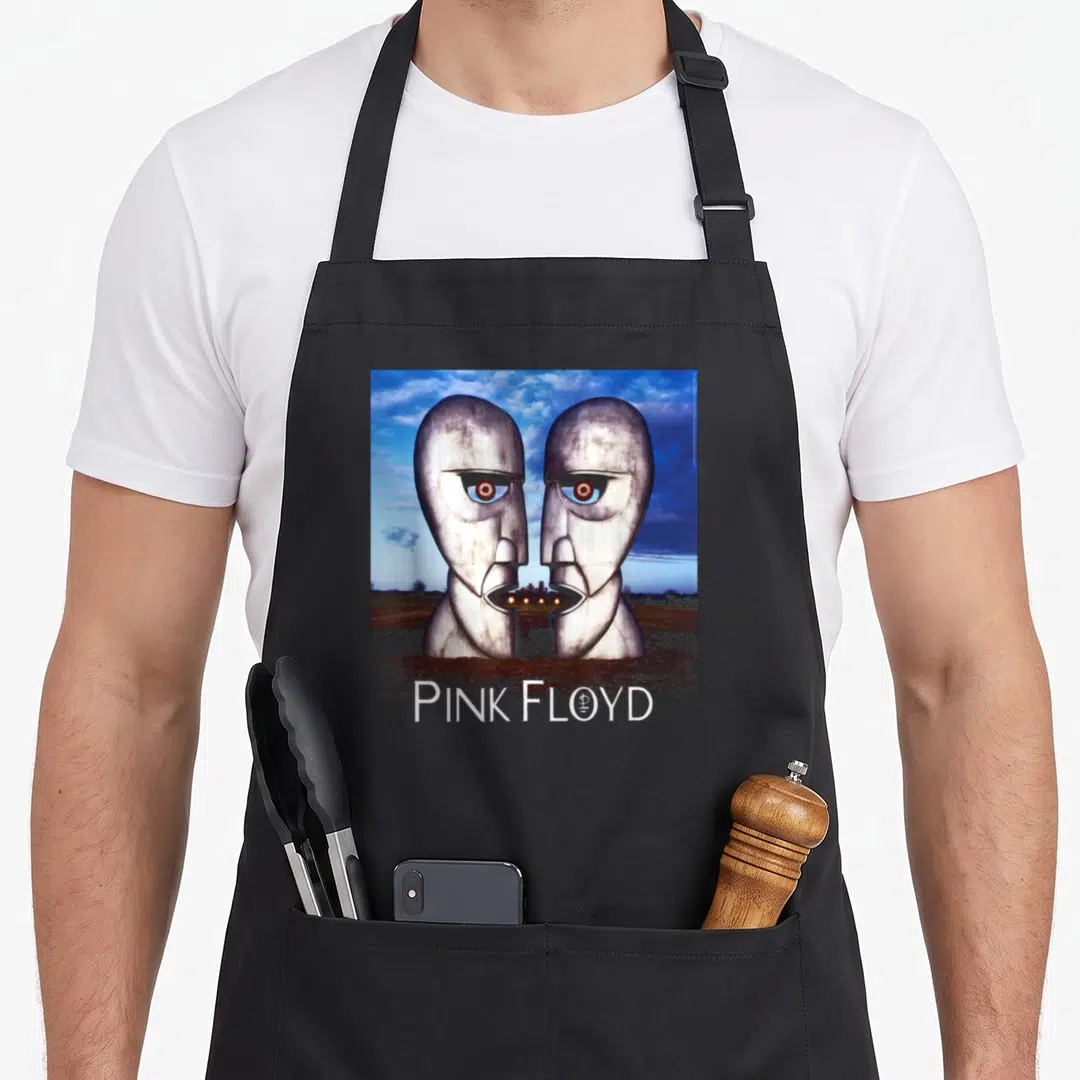 Floyd progressive apron - Two silver metal heads face each other over brown field under blue sky with PINK FLOYD text — shown on a black cotton canvas apron against a white background