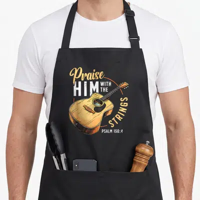 String bible christian apron - Retro acoustic guitar with Praise HIM WITH THE STRINGS text in orange and white — shown on a black cotton canvas apron against a white background