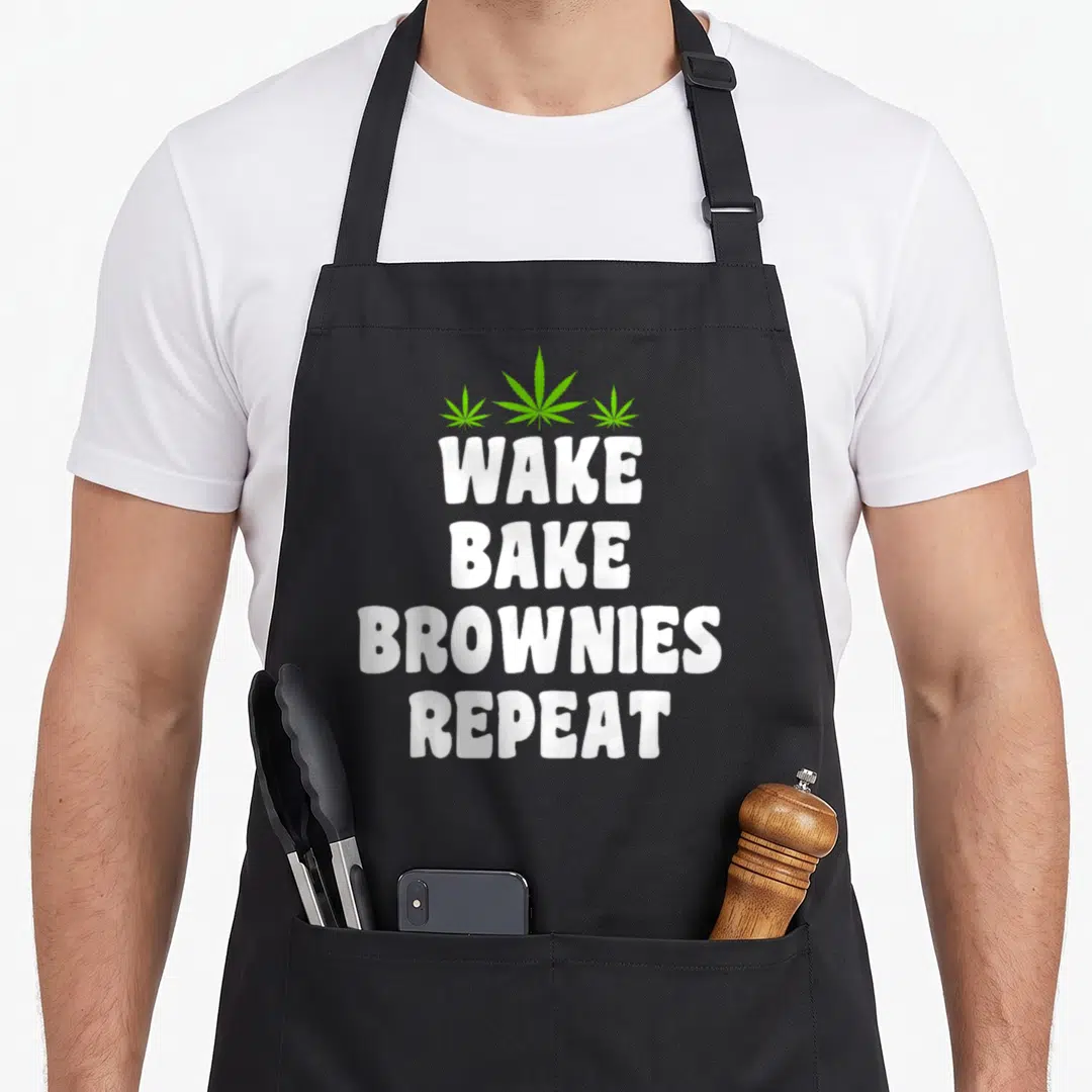 Browny smoker apron - Three green cannabis leaves above stacked white text reading WAKE BAKE BROWNIES REPEAT — shown on a black cotton canvas apron against a white background