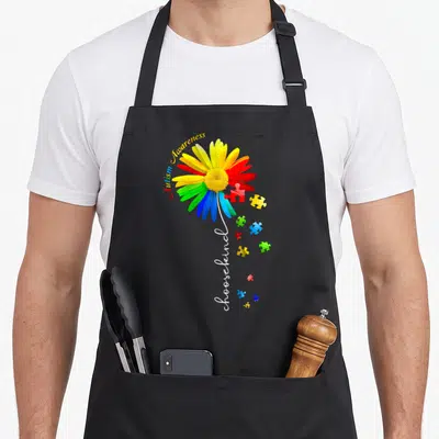 Choose sunflower apron - Rainbow daisy with scattered puzzle pieces and Autism Awareness choosekind text — shown on a black cotton canvas apron against a white background