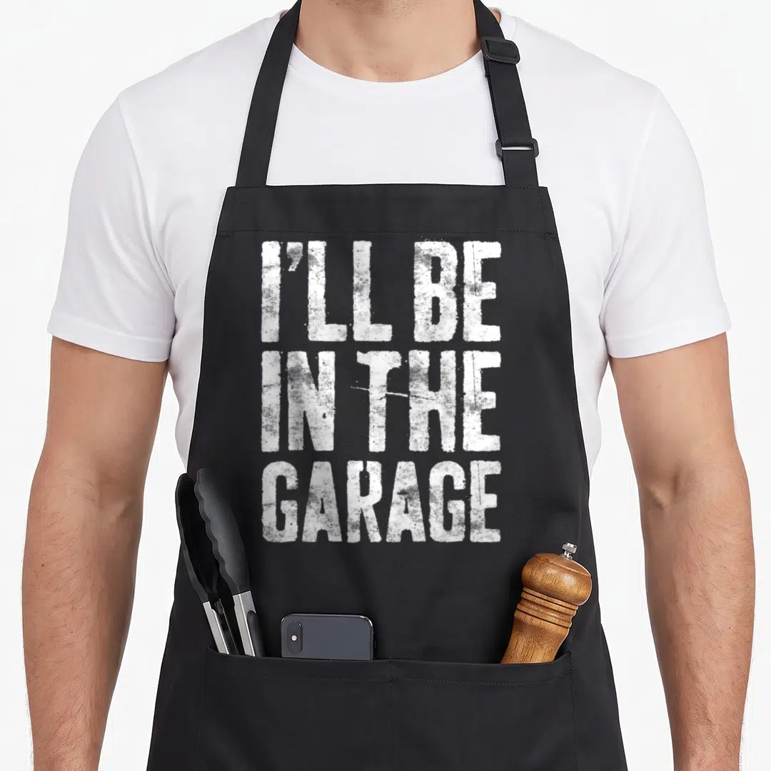 Garage handyman apron - White distressed text reading I'll Be In The Garage stacked in three lines on black fabric — shown on a black cotton canvas apron against a white background