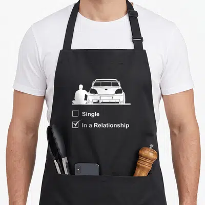 Culture design imports vintage apron - White graphic of man kneeling by car rear with single and in a relationship — shown on a black cotton canvas apron against a white background