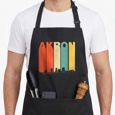 Akron 1970s apron - Tall colorful AKRON letters with grey skyline silhouette at the bottom — shown on a black cotton canvas apron against a white background