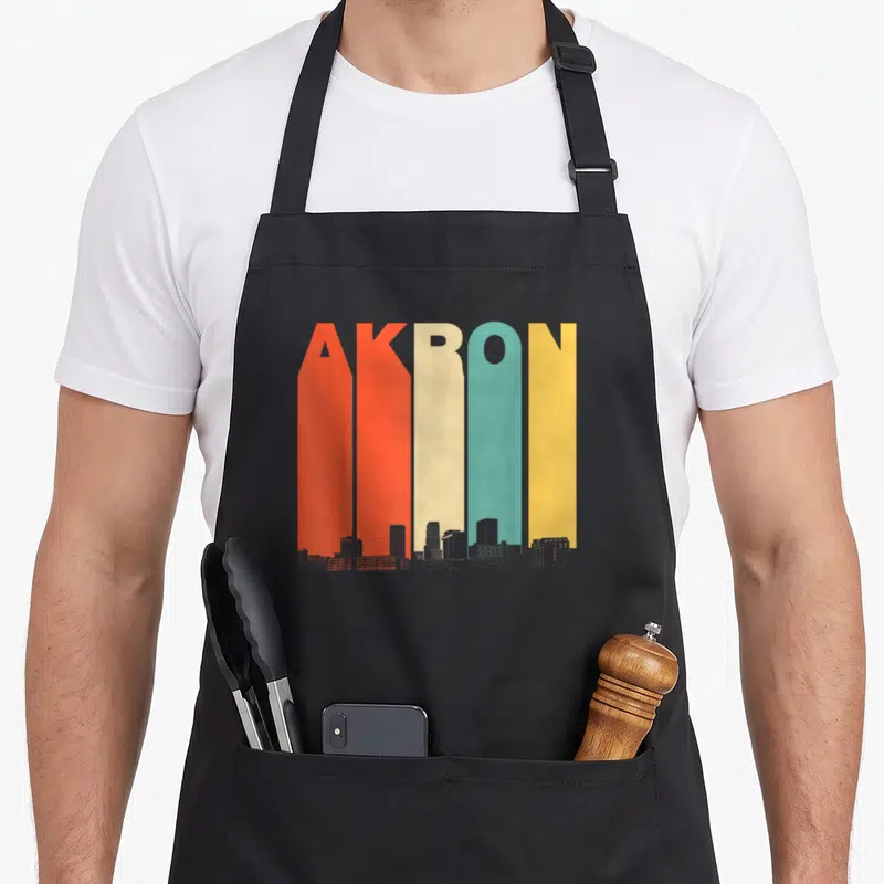 Akron 1970s apron - Tall colorful AKRON letters with grey skyline silhouette at the bottom — shown on a black cotton canvas apron against a white background