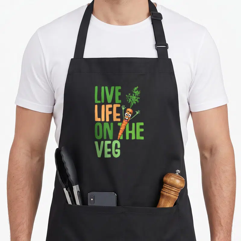 Live vegan apron - Stacked live life on the veg text with orange cartoon carrot overlapping letters — shown on a black cotton canvas apron against a white background