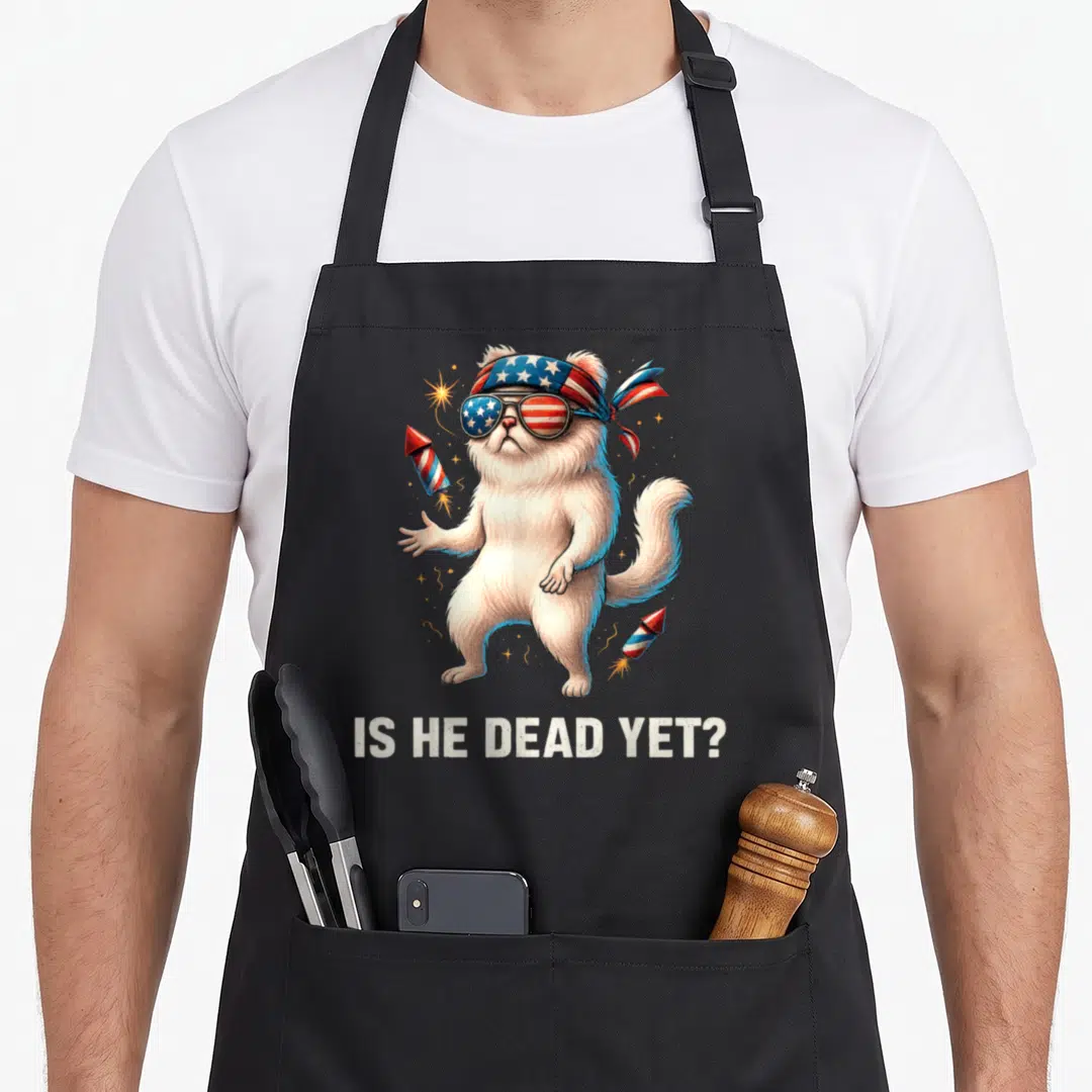 Dead yet cat apron - White cat with patriotic bandana and sunglasses holding fireworks below IS HE DEAD YET text — shown on a black cotton canvas apron against a white background