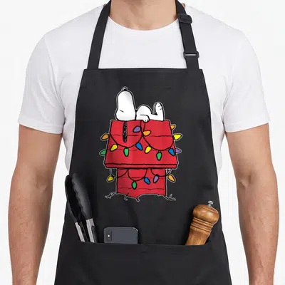 Doghouse christmas lights apron - Snoopy lying on red doghouse draped with yellow blue green and purple Christmas lights — shown on a black cotton canvas apron against a white background